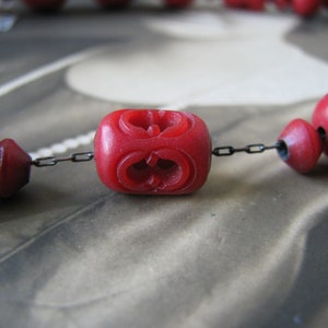 Peut inclure: Gros plan d'un collier de perles rouges avec une cha&icirc;ne. Les perles sont rondes et pr&eacute;sentent un motif sculpt&eacute;. Le collier est sur un fond blanc.
