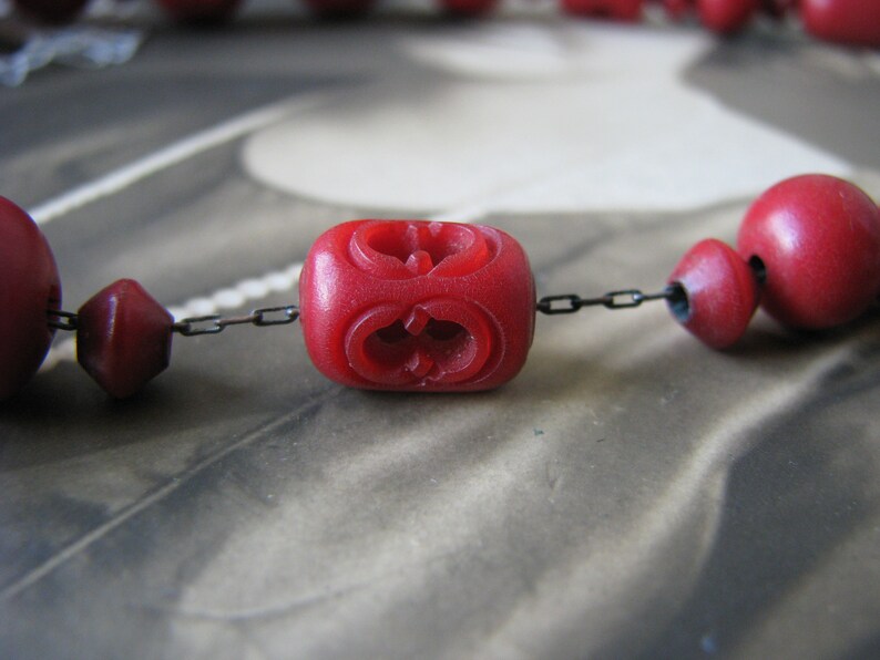 Peut inclure: Gros plan d'un collier de perles rouges avec une cha&icirc;ne. Les perles sont rondes et pr&eacute;sentent un motif sculpt&eacute;. Le collier est sur un fond blanc.
