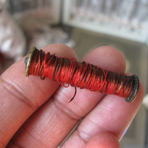 Puede incluir: Una pequeña bobina de hilo rojo con una tapa de metal dorado en cada extremo.