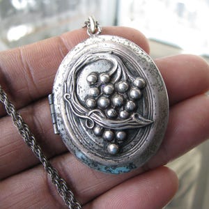 May include: Oval silver locket necklace with a detailed grape cluster design. The pendant is attached to a silver chain. The locket has a hinged opening and a tarnished, vintage appearance.