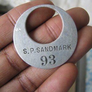 May include: A silver metal tag with the text "S.P. SANDMARK 93" engraved on it.