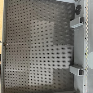 May include: A grey metal storage cabinet interior featuring a black pegboard back panel with a uniform grid of small circular holes. The cabinet includes two rectangular support brackets on the right side and a hinged lid mechanism at the top.
