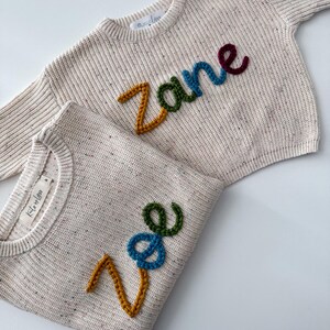 Peut inclure: Deux pulls en tricot cr&egrave;me avec des noms brod&eacute;s color&eacute;s. L'un des pulls porte le nom "Zane" en jaune, vert, bleu et bordeaux. L'autre porte le nom "Zoe" en jaune, bleu et vert. Les pulls ont une texture mouchet&eacute;e.