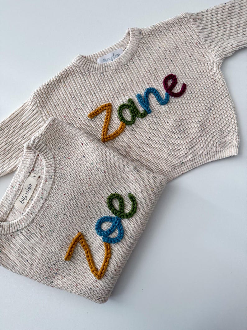 Peut inclure: Deux pulls en tricot cr&egrave;me avec des noms brod&eacute;s color&eacute;s. L'un des pulls porte le nom "Zane" en jaune, vert, bleu et bordeaux. L'autre porte le nom "Zoe" en jaune, bleu et vert. Les pulls ont une texture mouchet&eacute;e.