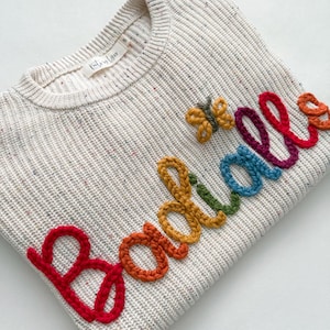 Peut inclure: Un pull cr&egrave;me avec un motif en tricot textur&eacute;. Le mot "Boodles" est brod&eacute; en couleurs arc-en-ciel sur le devant. Un petit papillon est cousu pr&egrave;s du mot. Le pull porte une &eacute;tiquette "Kathryn Lynn".