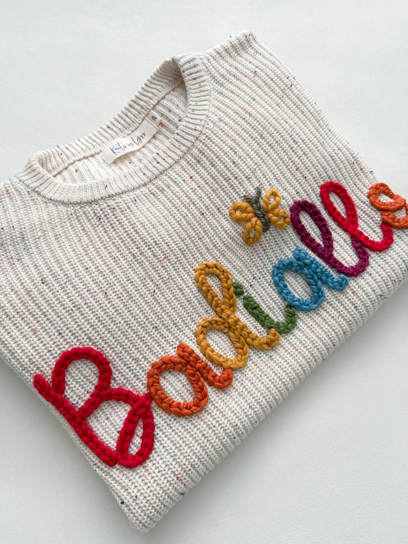 Peut inclure: Un pull cr&egrave;me avec un motif en tricot textur&eacute;. Le mot "Boodles" est brod&eacute; en couleurs arc-en-ciel sur le devant. Un petit papillon est cousu pr&egrave;s du mot. Le pull porte une &eacute;tiquette "Kathryn Lynn".