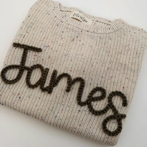 Peut inclure: Un pull en tricot beige avec le nom "James" brod&eacute; en fil brun fonc&eacute;. Le pull a un col rond et un motif mouchet&eacute; avec des &eacute;clats de couleur. L'&eacute;tiquette indique "Knits by Lyss".