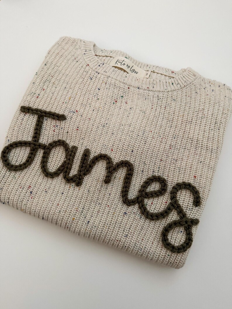 Peut inclure: Un pull en tricot beige avec le nom "James" brod&eacute; en fil brun fonc&eacute;. Le pull a un col rond et un motif mouchet&eacute; avec des &eacute;clats de couleur. L'&eacute;tiquette indique "Knits by Lyss".