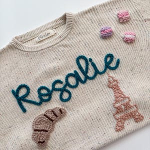 Peut inclure: Pull-over cr&egrave;me avec le nom "Rosalie" en script turquoise. Le pull pr&eacute;sente un croissant, la Tour Eiffel et trois macarons appliqu&eacute;s. Le pull a une texture mouchet&eacute;e et une petite &eacute;tiquette.