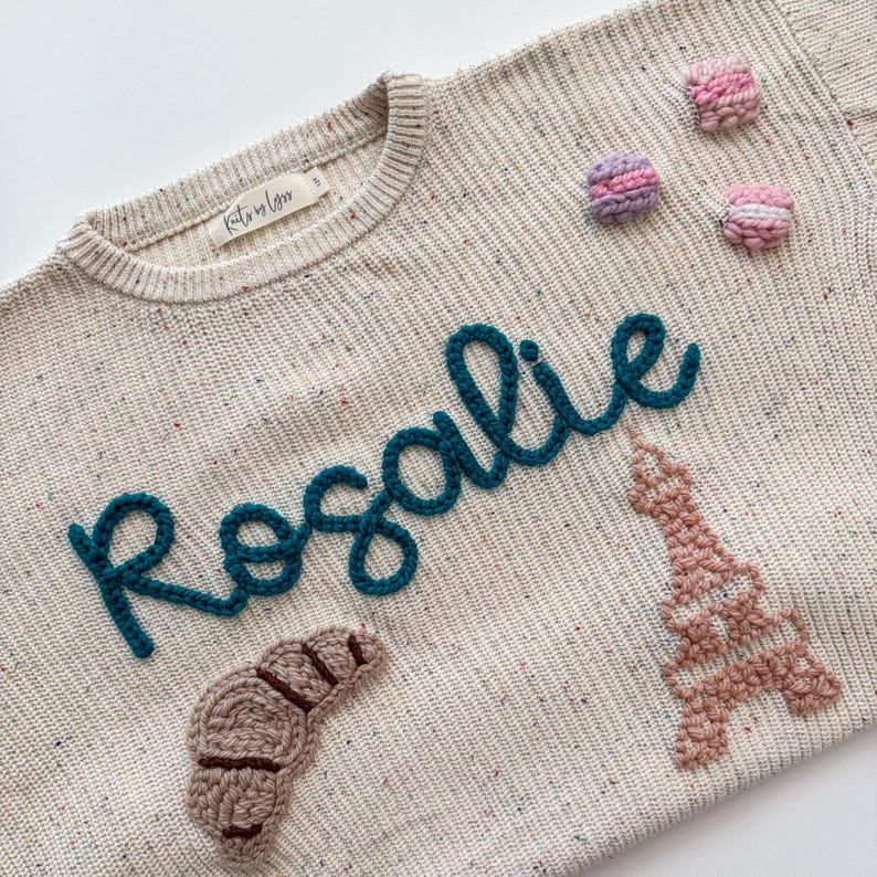 Peut inclure: Pull-over cr&egrave;me avec le nom "Rosalie" en script turquoise. Le pull pr&eacute;sente un croissant, la Tour Eiffel et trois macarons appliqu&eacute;s. Le pull a une texture mouchet&eacute;e et une petite &eacute;tiquette.