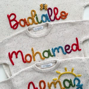 Peut inclure: Trois pulls cr&egrave;me avec des noms brod&eacute;s color&eacute;s : "Badiiallo", "Mohamed" et "Almaz". Chaque pull pr&eacute;sente un nom aux couleurs de l'arc-en-ciel, avec un petit papillon ou un soleil au-dessus des noms. Les pulls ont une texture mouchet&eacute;e.