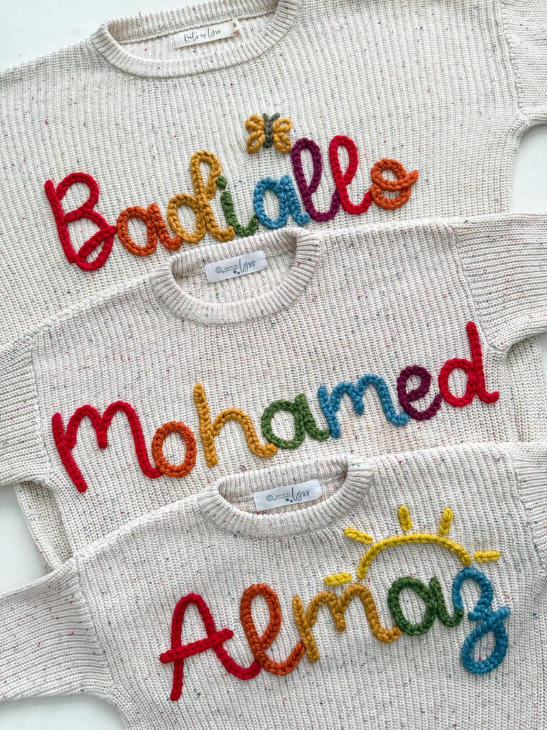 Peut inclure: Trois pulls cr&egrave;me avec des noms brod&eacute;s color&eacute;s : "Badiiallo", "Mohamed" et "Almaz". Chaque pull pr&eacute;sente un nom aux couleurs de l'arc-en-ciel, avec un petit papillon ou un soleil au-dessus des noms. Les pulls ont une texture mouchet&eacute;e.