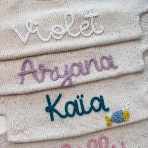 Peut inclure: Gros plan sur quatre pulls en tricot beige avec des noms brod&eacute;s : "Violet", "Aryana", "Kaia" et "Lilly". Le pull "Kaia" pr&eacute;sente un motif de poisson bleu et jaune. Les pulls ont un motif mouchet&eacute;.