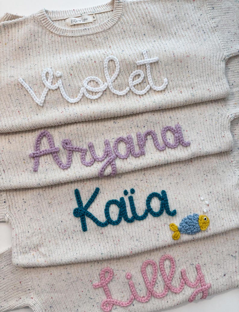 Peut inclure: Gros plan sur quatre pulls en tricot beige avec des noms brod&eacute;s : "Violet", "Aryana", "Kaia" et "Lilly". Le pull "Kaia" pr&eacute;sente un motif de poisson bleu et jaune. Les pulls ont un motif mouchet&eacute;.