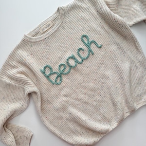 Peut inclure: Pull en tricot cr&egrave;me avec le mot "Beach" en script turquoise sur le devant. Le pull a un col rond et des manches longues. Le tissu tricot&eacute; a des mouchetures de couleur.