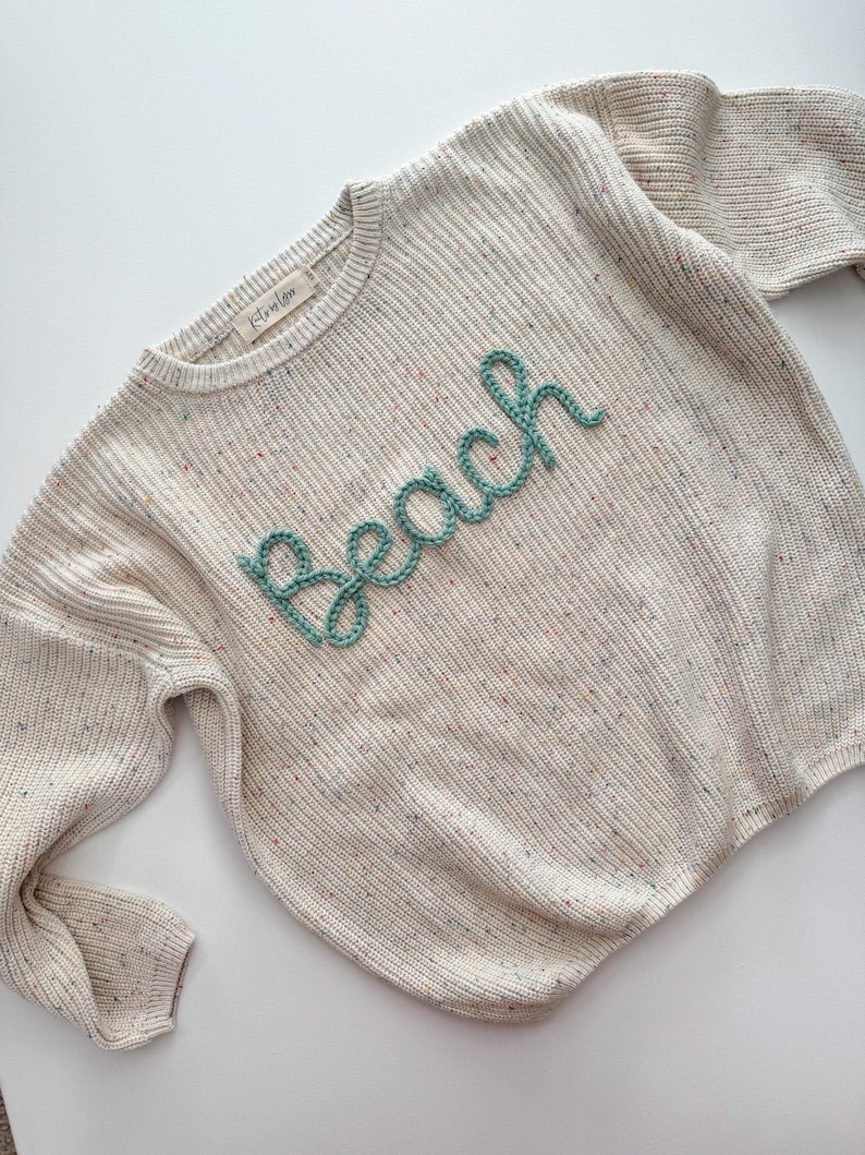 Peut inclure: Pull en tricot cr&egrave;me avec le mot "Beach" en script turquoise sur le devant. Le pull a un col rond et des manches longues. Le tissu tricot&eacute; a des mouchetures de couleur.