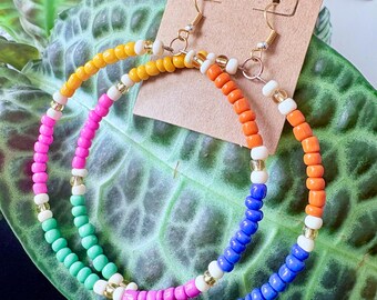 May include: Colorful beaded hoop earrings. One earring features yellow, orange, white, and pink beads. The other earring has orange, blue, white, and pink beads. The earrings are displayed on a brown card with gold-colored earring hooks.
