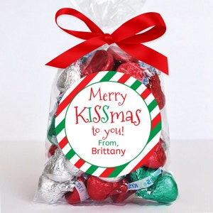 May include: A clear cellophane bag tied with a red ribbon, filled with foil-wrapped chocolate candies. A round sticker with the text "Merry KISSmas to you! From, Brittany" is attached to the bag. The candies are red, green, and silver.