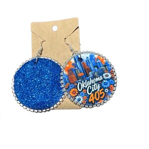 Op de afbeelding: Een paar ronde bungelende oorbellen met zilverkleurige metalen randen en ingebedde heldere strass-steentjes. De ene oorbel is bedekt met felblauwe glitters, terwijl de andere een afbeelding van de skyline van Oklahoma City toont met de tekst Oklahoma City 405.