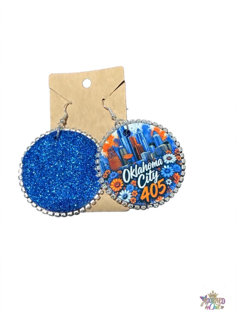 Op de afbeelding: Een paar ronde bungelende oorbellen met zilverkleurige metalen randen en ingebedde heldere strass-steentjes. De ene oorbel is bedekt met felblauwe glitters, terwijl de andere een afbeelding van de skyline van Oklahoma City toont met de tekst Oklahoma City 405.