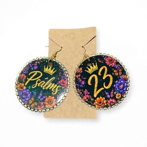 May include: A pair of round dangle earrings featuring a black background with colourful floral patterns and butterflies. The left earring displays the gold text Psalms and a crown, while the right earring shows the number 23 and a crown.