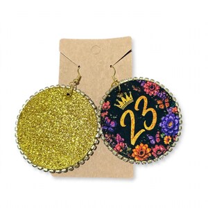 May include: A pair of round dangle earrings featuring a gold glitter finish on one side and a black background with a floral pattern, a gold crown icon, and the number 23 on the other, both bordered by small clear rhinestones on metal hooks.