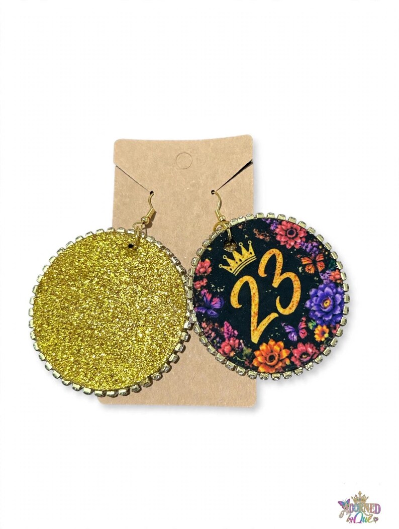 May include: A pair of round dangle earrings featuring a gold glitter finish on one side and a black background with a floral pattern, a gold crown icon, and the number 23 on the other, both bordered by small clear rhinestones on metal hooks.