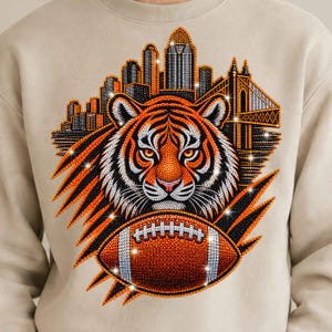 Op de afbeelding: Een beige sweater met ronde hals en een afbeelding van een tijgerkop, een football en een stadsskyline met een brug. Het ontwerp is opgebouwd uit kleine, sprankelende, veelkleurige stippen in de kleuren oranje, zwart, wit en zilver.