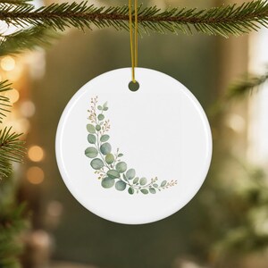 May include: A round white ceramic ornament featuring a delicate watercolor illustration of a curved eucalyptus branch with green leaves and small golden berries, suspended by a thin gold string for hanging on a tree branch.