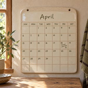 Puede incluir: Un calendario de pared de cerámica de color blanco roto y forma rectangular con acabado moteado. Presenta el nombre del mes April en la parte superior, seguido de una cuadrícula de días de domingo a sábado. El día 18 incluye el texto Dinner with Sam.