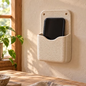 Könnte beinhalten: Eine wandmontierte, cremefarbene Ablage mit Sprenkeln, in der ein schwarzes Smartphone steckt. Der rechteckige Halter verfügt über eine geschwungene Öffnung an der Vorderseite und zwei runde Befestigungslöcher an der Oberseite.