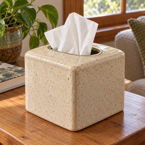May include: A square tissue box cover with a glossy, cream-colored ceramic finish featuring small brown speckles throughout. The box has a smooth, rounded edge design and a central rectangular opening at the top, from which white tissues emerge.