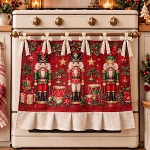 Op de afbeelding: Een rode ovenhoes met drie notenkrakerfiguren, trommels en kerstversieringen. De stof is versierd met hulst en sterren, afgewerkt met zes crèmekleurige kanten strikken aan de bovenkant en een geplooide crèmekleurige stoffen rand aan de onderkant.