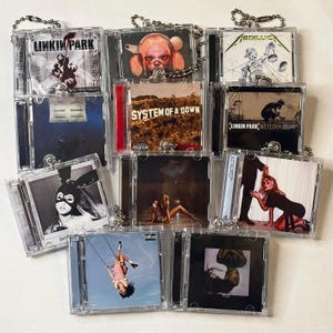 May include: A collection of miniature clear plastic CD cases featuring various album cover art, including Linkin Park, Metallica, System of a Down, and Ariana Grande. Each case is attached to a silver metal ball chain, creating a set of music-themed charms.