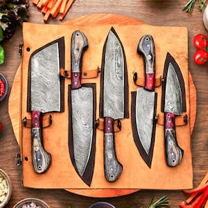May include: A set of five Damascus steel kitchen knives with dark wood and red handles, each housed in a custom leather sheath, arranged on a leather roll-up mat surrounded by fresh vegetables, herbs, and small bowls of ingredients.