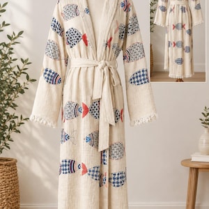 May include: A long-sleeved, floor-length robe in a light beige color featuring a repeating pattern of stylized fish in blue, red, and black. The robe includes a matching tie belt at the waist, a hood, and frayed fringe detailing along the hem.