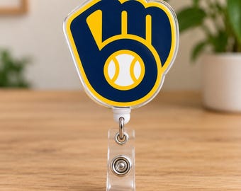 Puede incluir: Un portacredenciales retráctil con el logotipo de los Milwaukee Brewers, que consiste en un guante de béisbol azul marino que forma las letras M y B con una pelota de béisbol blanca en el centro, todo delineado en amarillo sobre una base de plástico transparente.
