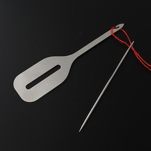 May include: A silver-toned metal needle threader with a flat, rectangular handle featuring a central slot, paired with a long, thin sewing needle threaded with a single strand of bright red thread.