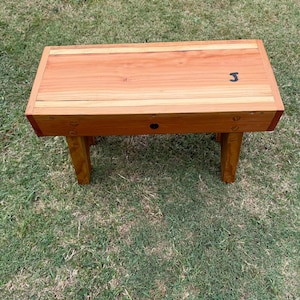 May include: A rectangular wooden bench featuring a light brown grain pattern on the top surface and a darker brown finish on the four legs. The top surface displays a small black letter J printed on the right side and a single circular hole.