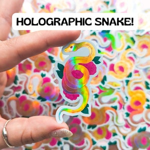 May include: A holographic sticker of a yellow snake with pink flowers. The sticker is iridescent and has a rainbow sheen. The sticker is being held by a hand.