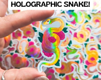 May include: A holographic sticker of a yellow snake with pink flowers. The sticker is iridescent and has a rainbow sheen. The sticker is being held by a hand.