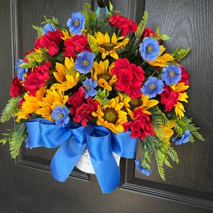 以下が含まれることがあります： ヒマワリ、赤いバラ、青い花が特徴の鮮やかな花のリース。緑の葉と明るい青のリボンがアクセントになっています。リースは黒いドアに飾られています。