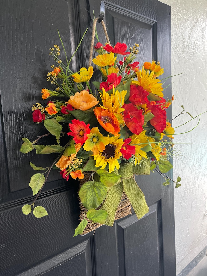 以下が含まれることがあります： ひまわり、ポピー、その他の黄色、オレンジ、赤色のカラフルな花々が咲き誇る、編みかごに入った活気あふれるフラワーアレンジメント。緑のリボンが装飾的なアクセントを添えています。かごはロープで吊るされています。