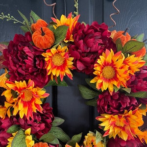 May include: A fall-themed wreath with burgundy peonies, orange and yellow sunflowers, and small orange pumpkins. The wreath is made of artificial flowers and is perfect for decorating a door or wall.