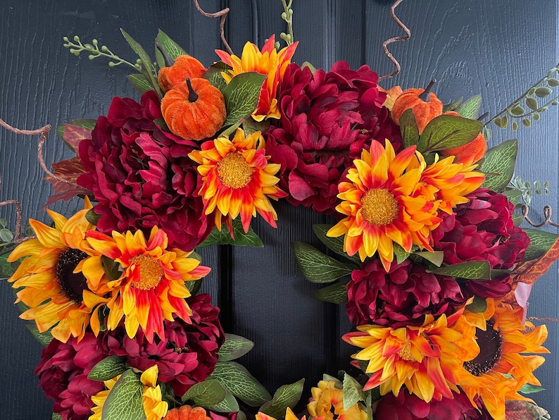 May include: A fall-themed wreath with burgundy peonies, orange and yellow sunflowers, and small orange pumpkins. The wreath is made of artificial flowers and is perfect for decorating a door or wall.