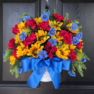 以下が含まれることがあります： ヒマワリ、赤いバラ、青い花が特徴の鮮やかな花のリース。緑の葉と大きな青いリボンがアクセントになっています。リースは黒いドアに飾られています。