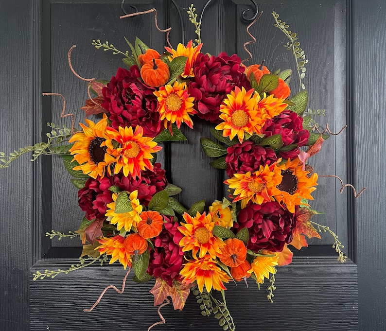 May include: A fall-themed wreath with burgundy, orange, and yellow flowers. The wreath is decorated with green leaves and small pumpkins.