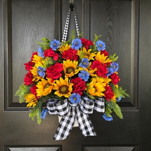 以下が含まれることがあります： ヒマワリ、赤いバラ、青い花が特徴の鮮やかな花のリース。緑の葉と白黒のチェック柄のリボンがアクセントになっています。黒いドアから吊り下げられています。