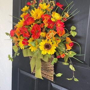 以下が含まれることがあります： ヒマワリ、ポピー、その他のカラフルな花々が、黄色、オレンジ、赤の色合いで飾られた活気のあるフラワーバスケットアレンジメント。 バスケットは織り込まれた小枝で作られ、緑のリボンで飾られています。 ジュートロープから吊り下げられています。