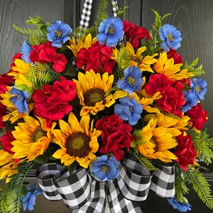 以下が含まれることがあります： ヒマワリ、赤いバラ、青い花が特徴の鮮やかな花のリース。緑の葉を含み、白黒のチェック柄のリボンでアクセントを加えています。ドアの装飾に最適です。